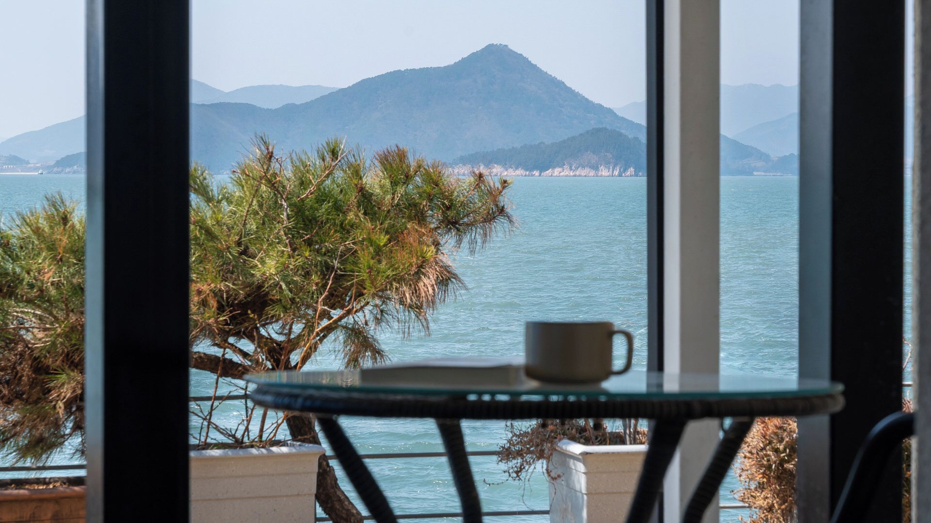 A view of a coastal landscape with a mountain in the distance, a table with a cup, and a chair.