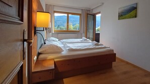Un dormitorio con una cama de madera, una lámpara de noche, una ventana con vistas a montañas y un cuadro en la pared.