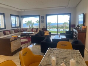 A spacious living room with a sofa set, a dining table, and a view of the ocean. - Villa Deraoui (Tangier-Tétouan-Al Hoceima)