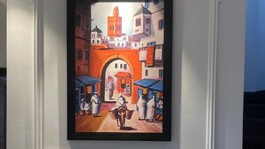 Un tableau encadré représentant une rue animée, avec une arche, est accroché à un mur blanc orné de moulures décoratives.