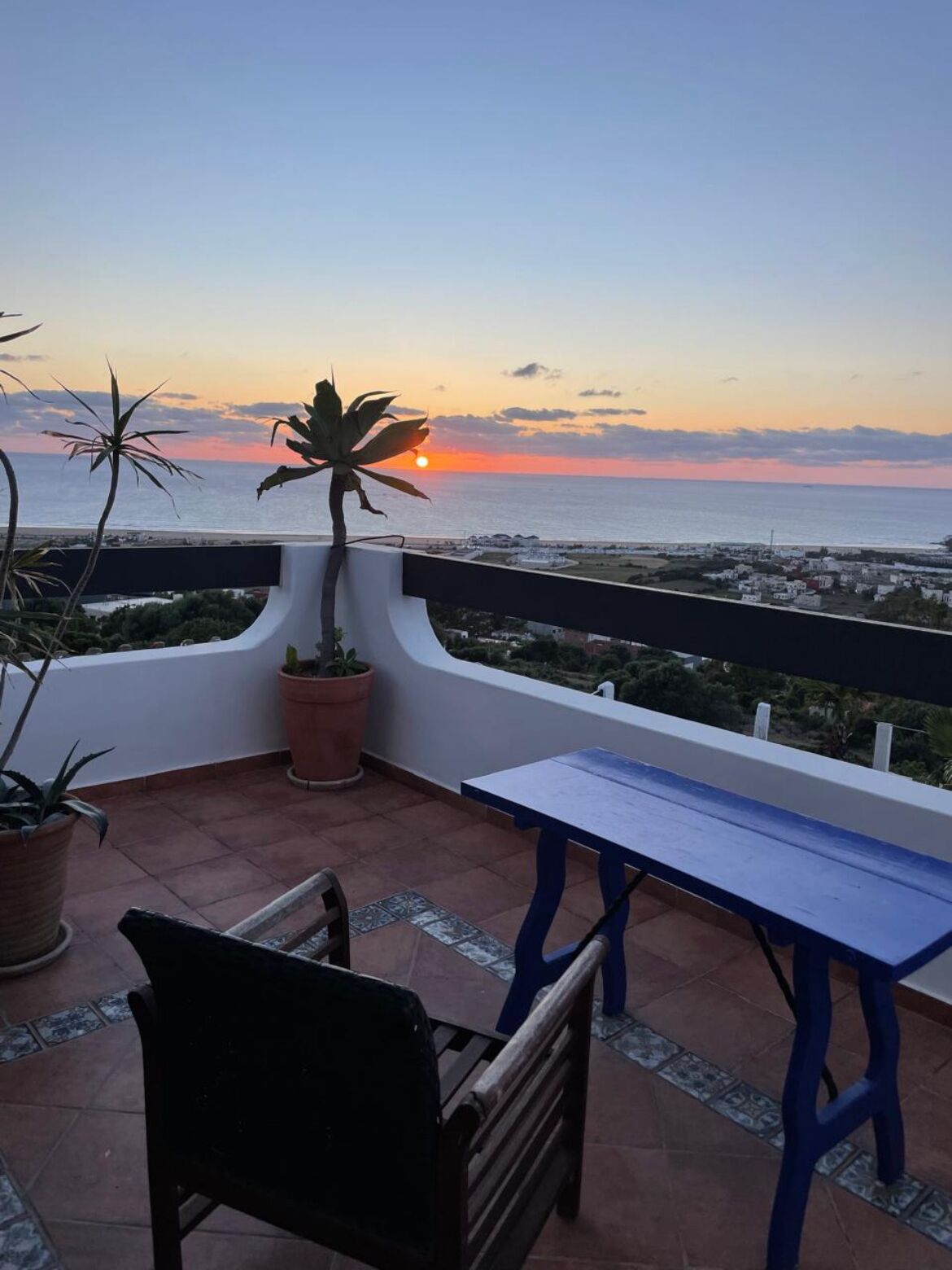 A terrace with a view of the sunset over the sea, a potted plant, and a table and chair.
