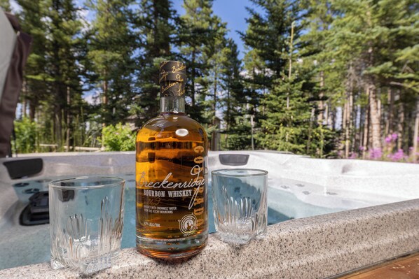 A bottle of bourbon whiskey and two glasses on a stone ledge by a hot tub surrounded by trees.