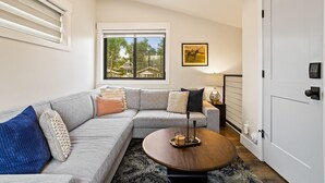 En moderne stue med en modulsofa, et rundt sofabord, et sengebord med lampe og et stort vindue med udsigt til huse.