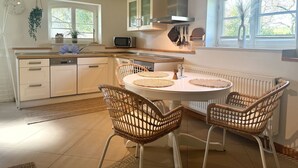A modern kitchen with white cabinetry, a round dining table with two wicker chairs, a microwave, and a hanging plant. - Ferienwohnung Alvine - Dubnitz - Doppelhaushälfte Alwine (Sassnitz)