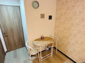 Quarto pequeno com porta de madeira, um relógio na parede, uma mesa com cadeiras e papel de parede estampado.