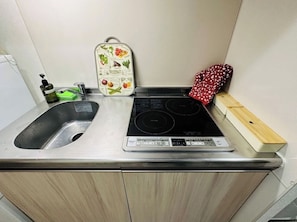 Uma cozinha com pia de aço inoxidável, fogão por indução preto, tábua de corte de madeira e um frasco verde de detergente.