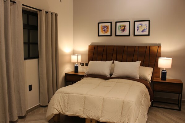 Una habitación de hotel con una cama grande, dos mesitas de noche con lámparas, una ventana con cortinas y tres cuadros enmarcados en la pared.