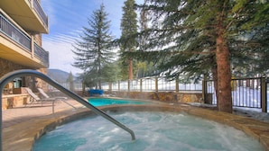 A hot tub with a metal ladder, surrounded by trees and a stone wall, with a pool in the background. - 3br Ski-in/ski-out~on Mountain~lone Eagle 3053 3 Bedroom Condo by RedAwning (Keystone)