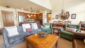 A spacious living room with a grey sofa, a wooden coffee table, and a green armchair. A kitchen with wooden cabinets and stainless steel appliances is visible in the background. - 3br Ski-in/ski-out~on Mountain~lone Eagle 3053 3 Bedroom Condo by RedAwning (Keystone)