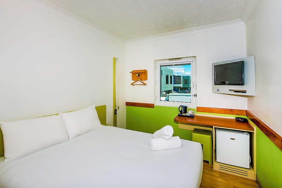 Hotellihuone, jossa on sänky, pieni työpöytä, televisio ja ikkuna, josta on näkymä rakennuksiin.