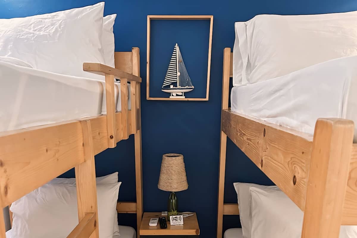 Et lille værelse med to køjesenge i træ, et lille bord med en lampe og et billede i ramme på væggen.