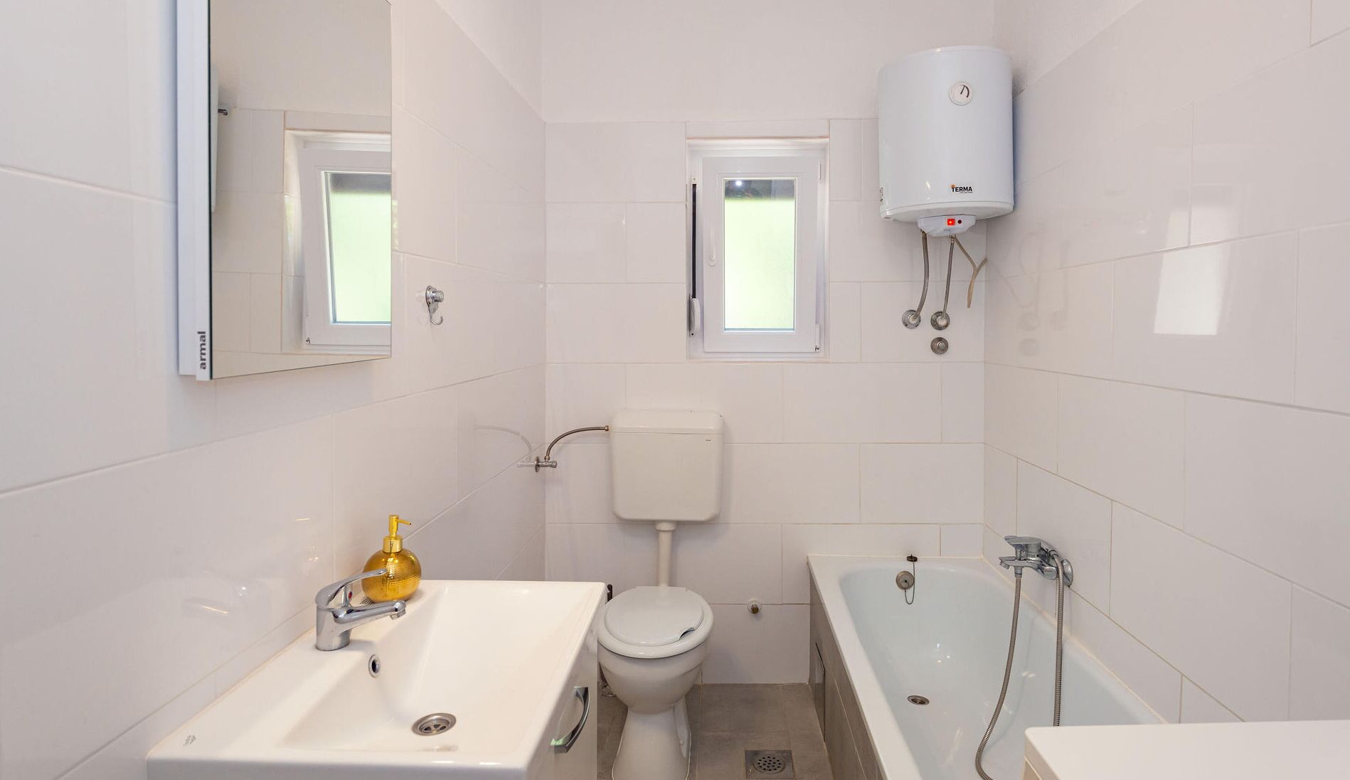 A small, modern bathroom with white tiles, a white sink, a toilet, a bathtub, and a wall-mounted water heater.