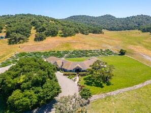 An aerial view of a large estate with a central building surrounded by expansive green lawns and a hilly backdrop.