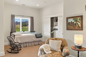 Quarto com cama, poltrona de balanço, cadeira, mesa de cabeceira com abajur e um quadro na parede.