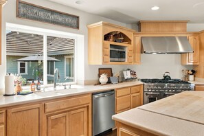 A kitchen with wooden cabinets, a stainless steel range hood, a dishwasher, and a window with a view of a house.