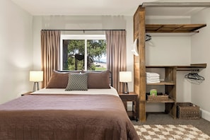 Un dormitorio con cama, dos lámparas de noche, una ventana con cortinas, un estante de madera con toallas y una cesta de mimbre.
