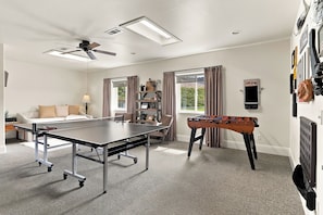 Una habitación con mesa de ping pong, mesa de futbolín, ventilador de techo y ventana con cortinas.