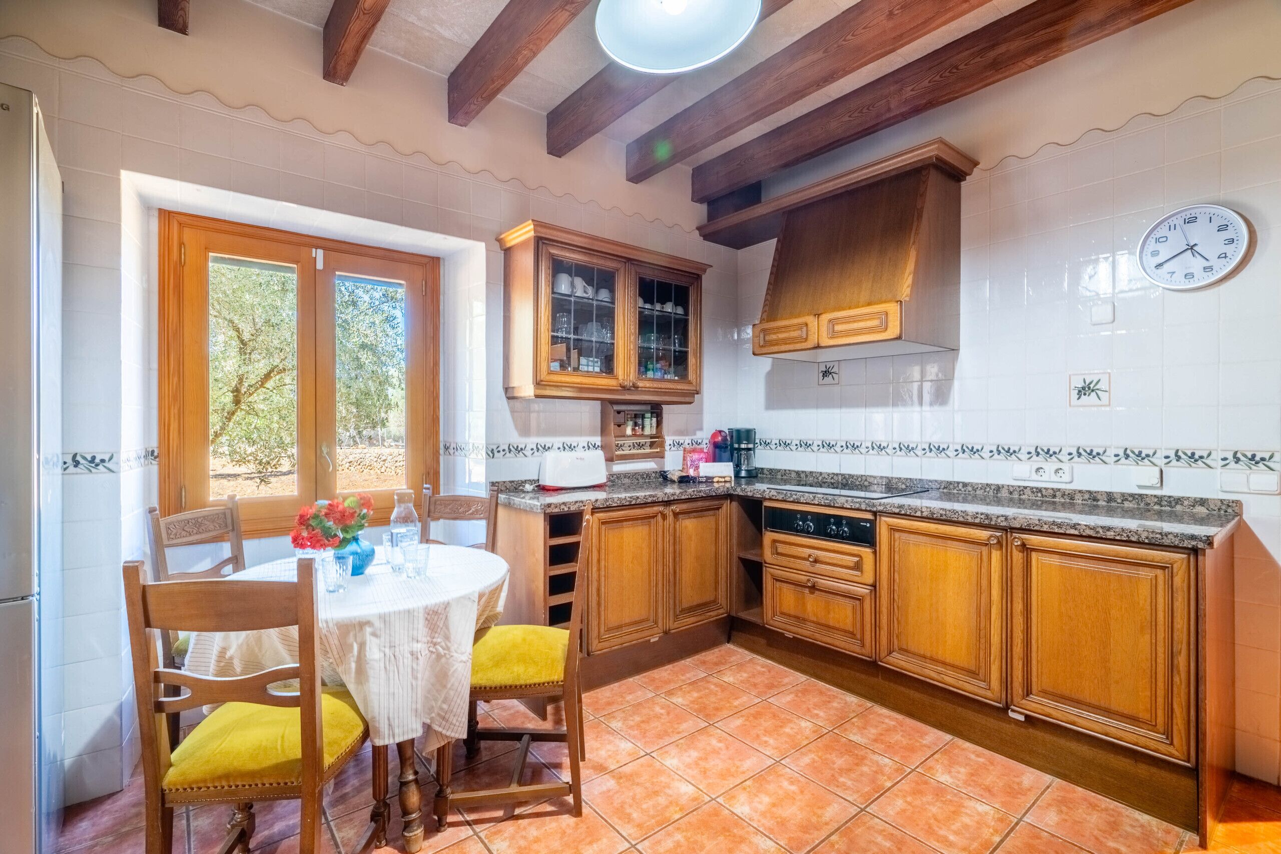 Una cocina con armarios de madera, salpicadero de azulejos blancos, un reloj y un comedor con mesa y sillas.