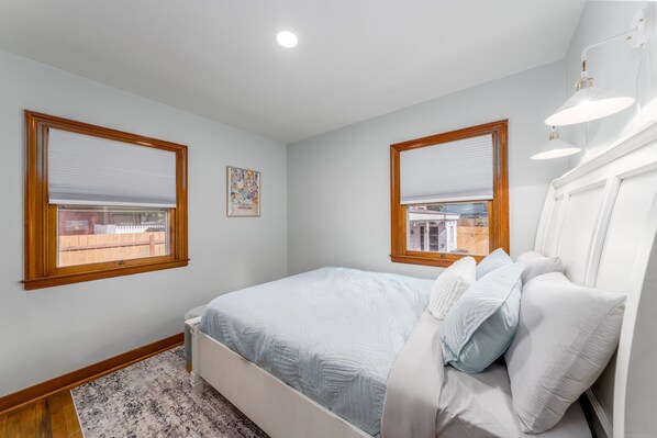 Une chambre à coucher avec un lit, deux fenêtres équipées de stores, un luminaire au plafond et un tapis.