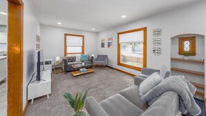 Un salon moderne avec un canapé gris, une table basse et un téléviseur. Il y a deux fenêtres équipées de stores, un plancher en bois et un mur blanc orné d’objets décoratifs.
