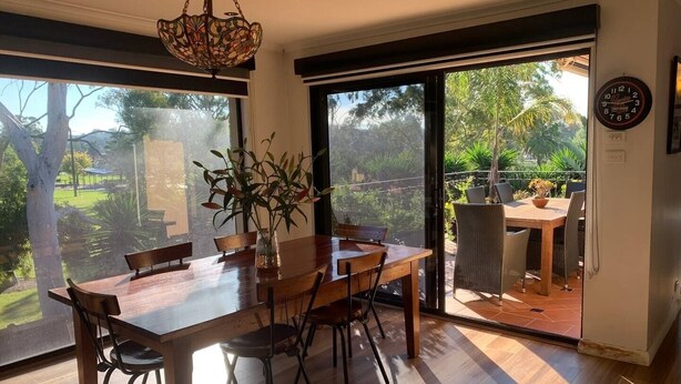 A dining area with a wooden table and chairs, a vase with plants, and a view of a garden through large sliding doors. - Brum's Place | Inner City Tropical Haven (Maribyrnong)