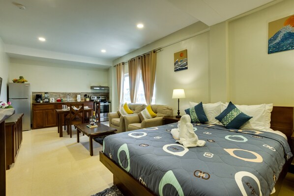 A hotel room with a bed, a desk, a chair, a sofa, a kitchen area, and a painting on the wall.