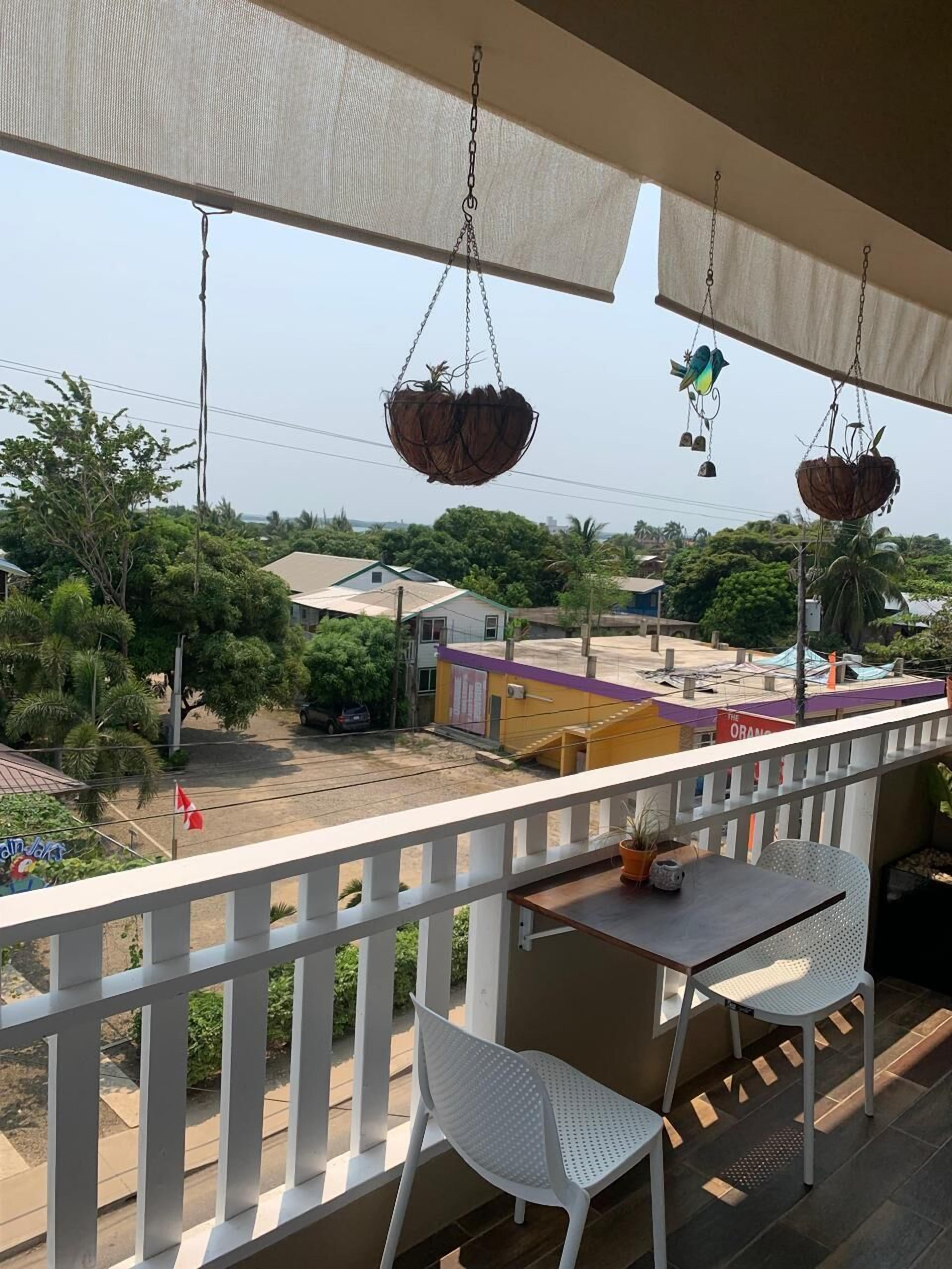 A balcony with white chairs, hanging baskets, and a view of a street with buildings and trees.