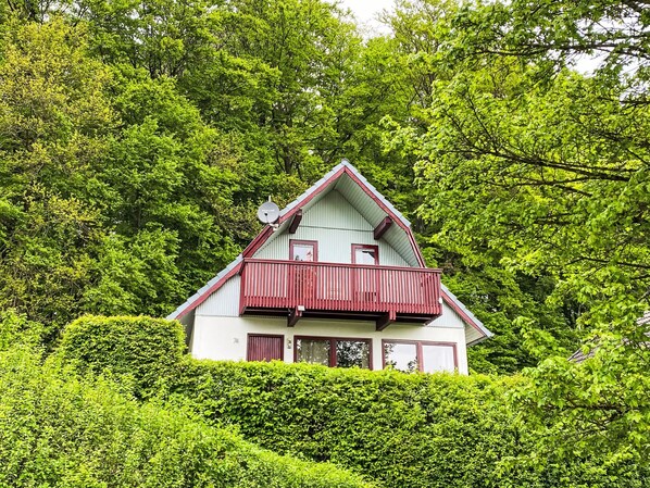 Een tweelaags huis met een rood balkon, omringd door weelderige groene begroeiing.