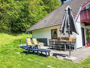 Een huis met een pannendak, een terras met tafel en stoelen, en een schommelset.