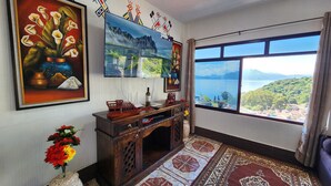 Ein Zimmer mit großem Fenster, das einen malerischen Blick auf einen See und Berge bietet, ein Holztischchen mit Dekoration, eine Vase mit Blumen und Wandbilder, darunter ein Blumenbild und ein Wandgemälde mit farbenfrohen, abstrakten Figuren.