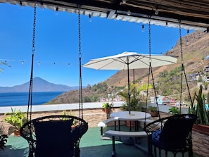 Une terrasse agrémentée de fauteuils suspendus, d’une table et d’un parasol, offrant une vue imprenable sur la côte, avec des maisons et une montagne en toile de fond.
