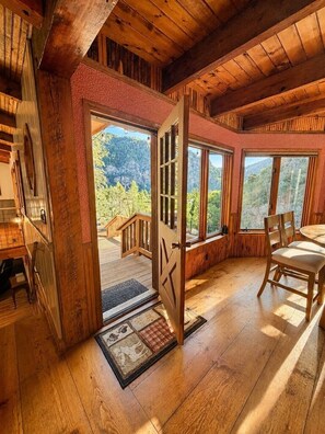 Un intérieur en bois avec une grande fenêtre, une porte donnant sur l’extérieur et un coin repas comprenant une table et des chaises.