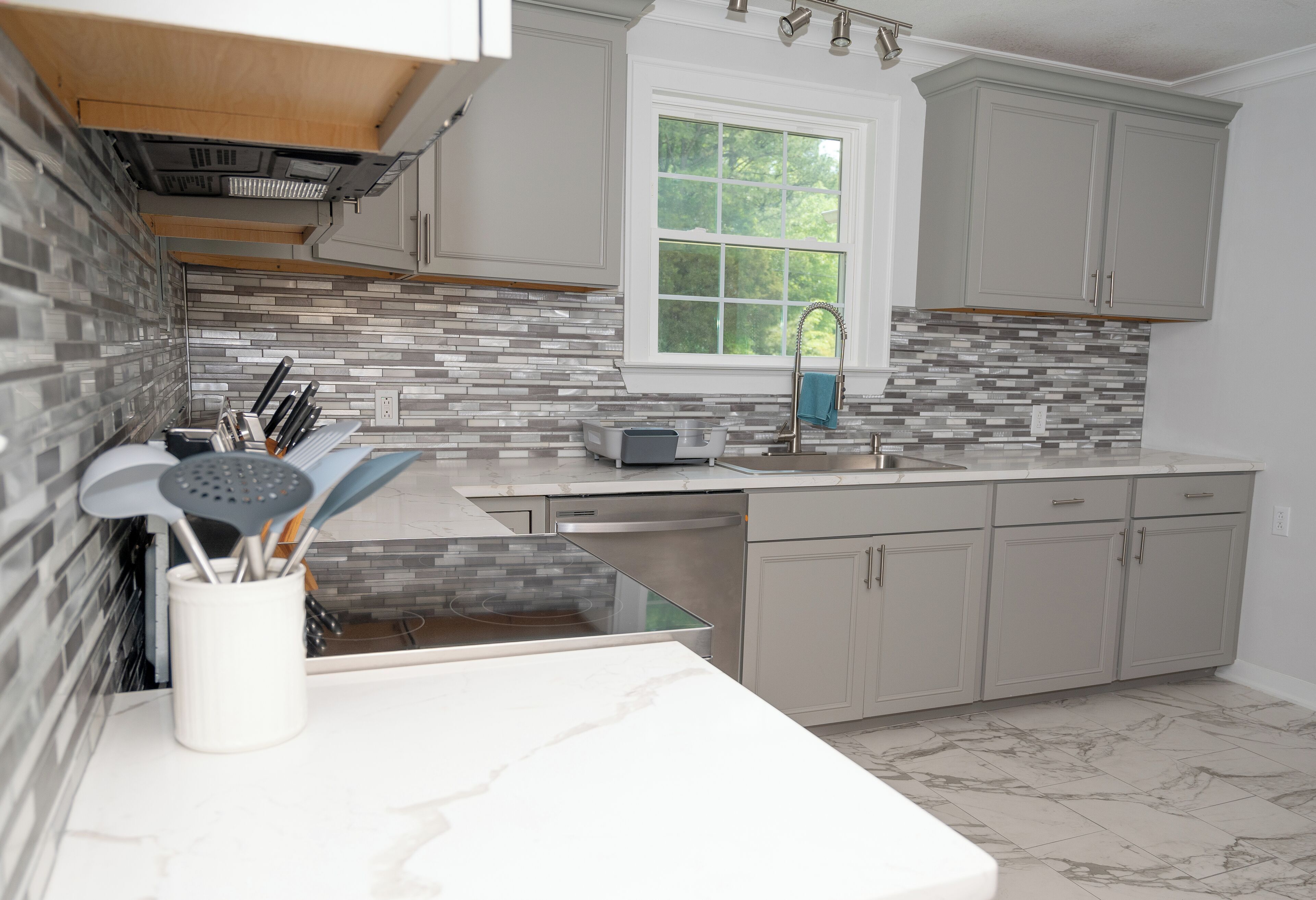 A modern kitchen with grey cabinets, a tiled backsplash, a window, and a dishwasher.