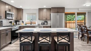 A modern kitchen with wooden cabinets, a central island with bar stools, stainless steel appliances, and a view of a backyard. - Perch - Spacious Modern Cabin w/ Hot Tub, BBQ & Cali- King-Sized Bed | Pet-Friendly (Ashford)
