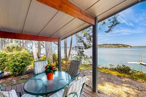 A covered outdoor seating area with a glass table and chairs, overlooking a lake with a bridge in the distance. - Family-friendly, soundfront getaway with deck, Wifi, W/D and mountain views (Fox Island)
