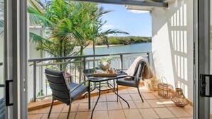 A balcony with a table, chairs, a potted plant, and a view of a lake and palm trees. - Waterfront at Sunrise Cove (Kingscliff)