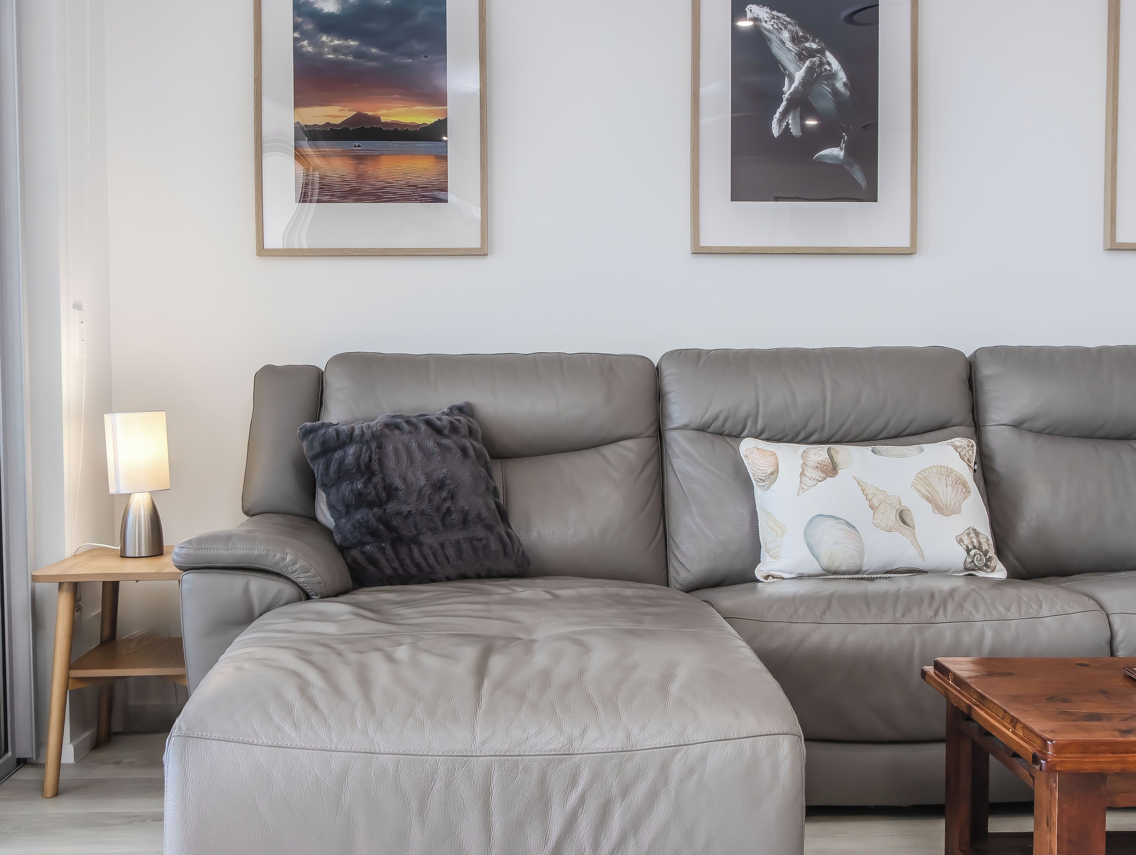 A modern living room with a grey leather sofa, a wooden coffee table, and a side table with a lamp. There are two framed pictures on the wall.