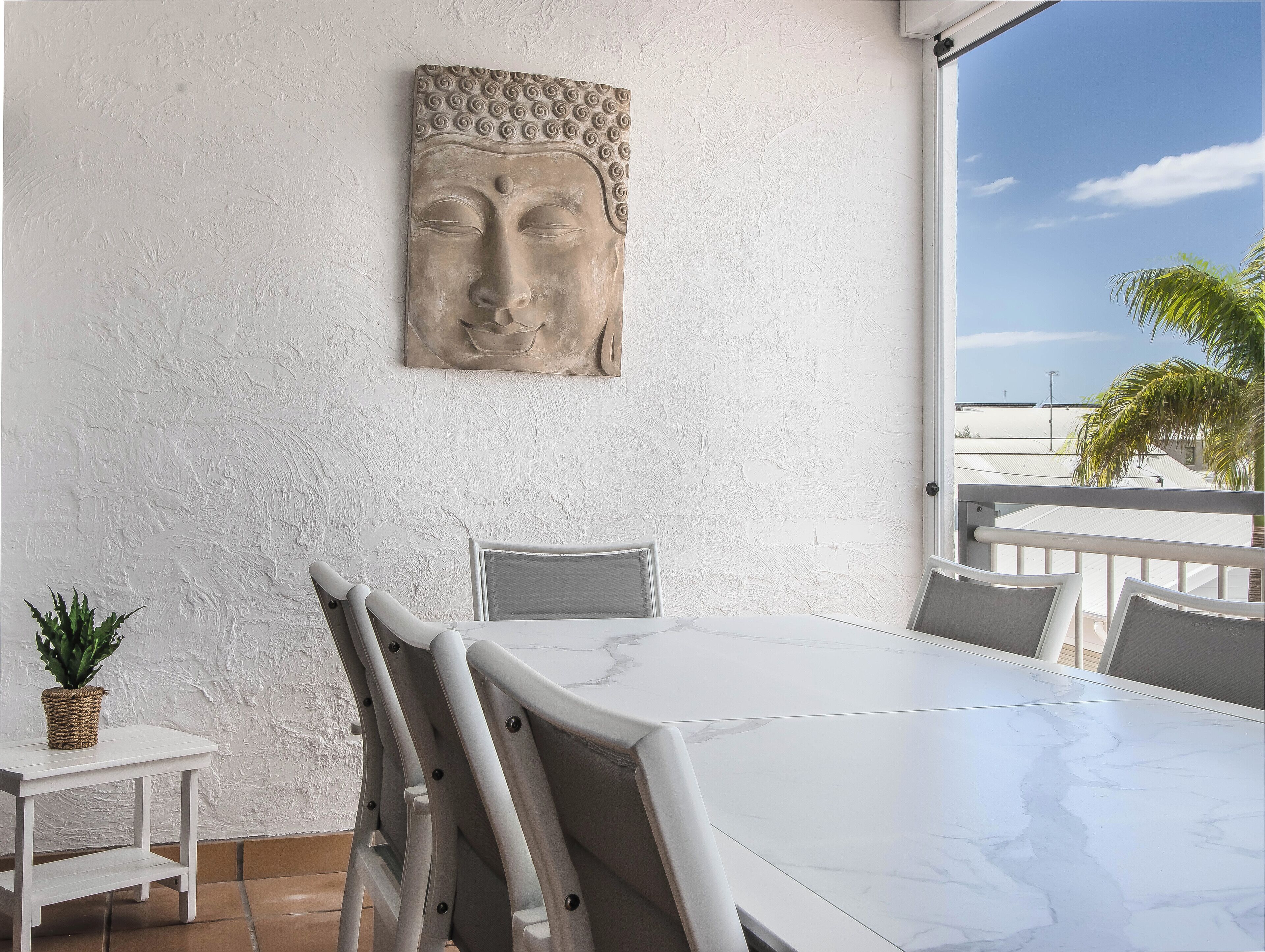 Ein Essbereich mit weißem Tisch und Stühlen, einer Buddha-Kopf-Skulptur an der Wand und Blick auf den Himmel und eine Palme.