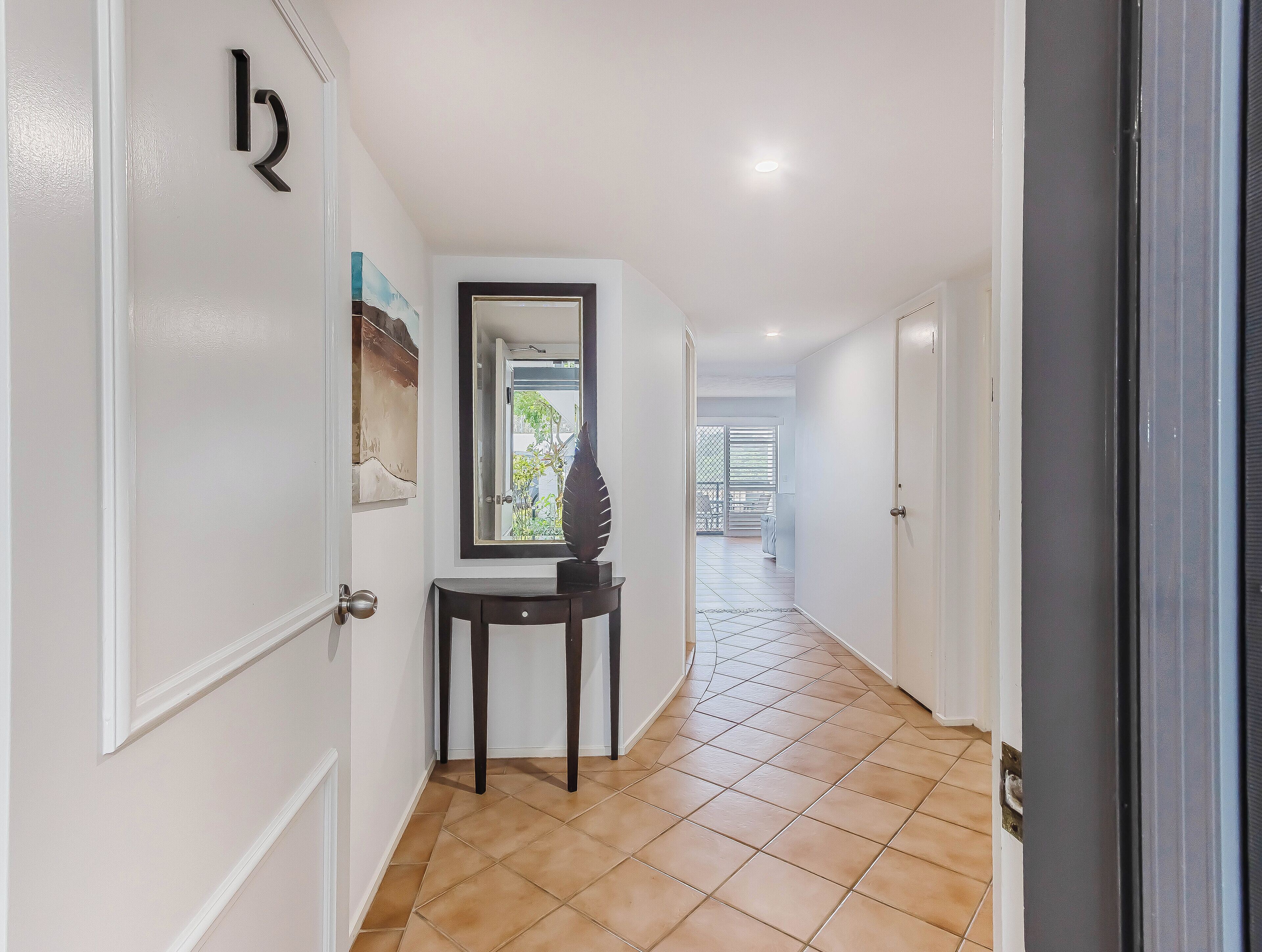 Un pasillo con piso de baldosas, una puerta con placa de número, una pequeña mesa con un objeto decorativo y una ventana con vista al exterior.