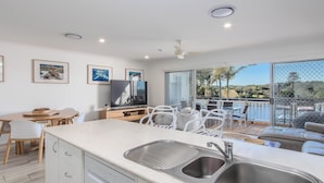 A modern kitchen with a built-in dishwasher, a dining area with white chairs, and a large TV. - Waterfront at Sunrise Cove (Kingscliff)