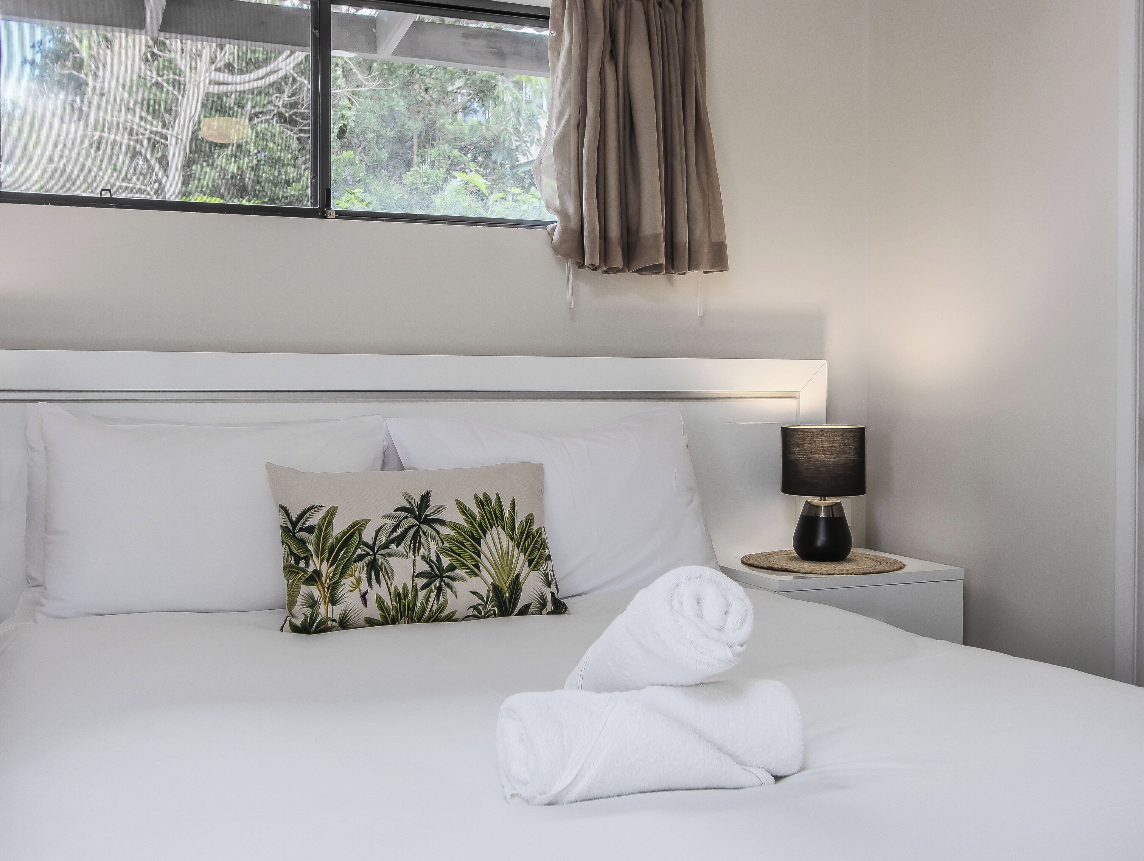 A neatly made bed with white linens, a tropical leaf pillow, and rolled towels on the bed.