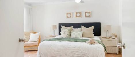 A bedroom with a bed, a nightstand, a chair, a ceiling fan, and a wall-mounted light fixture.