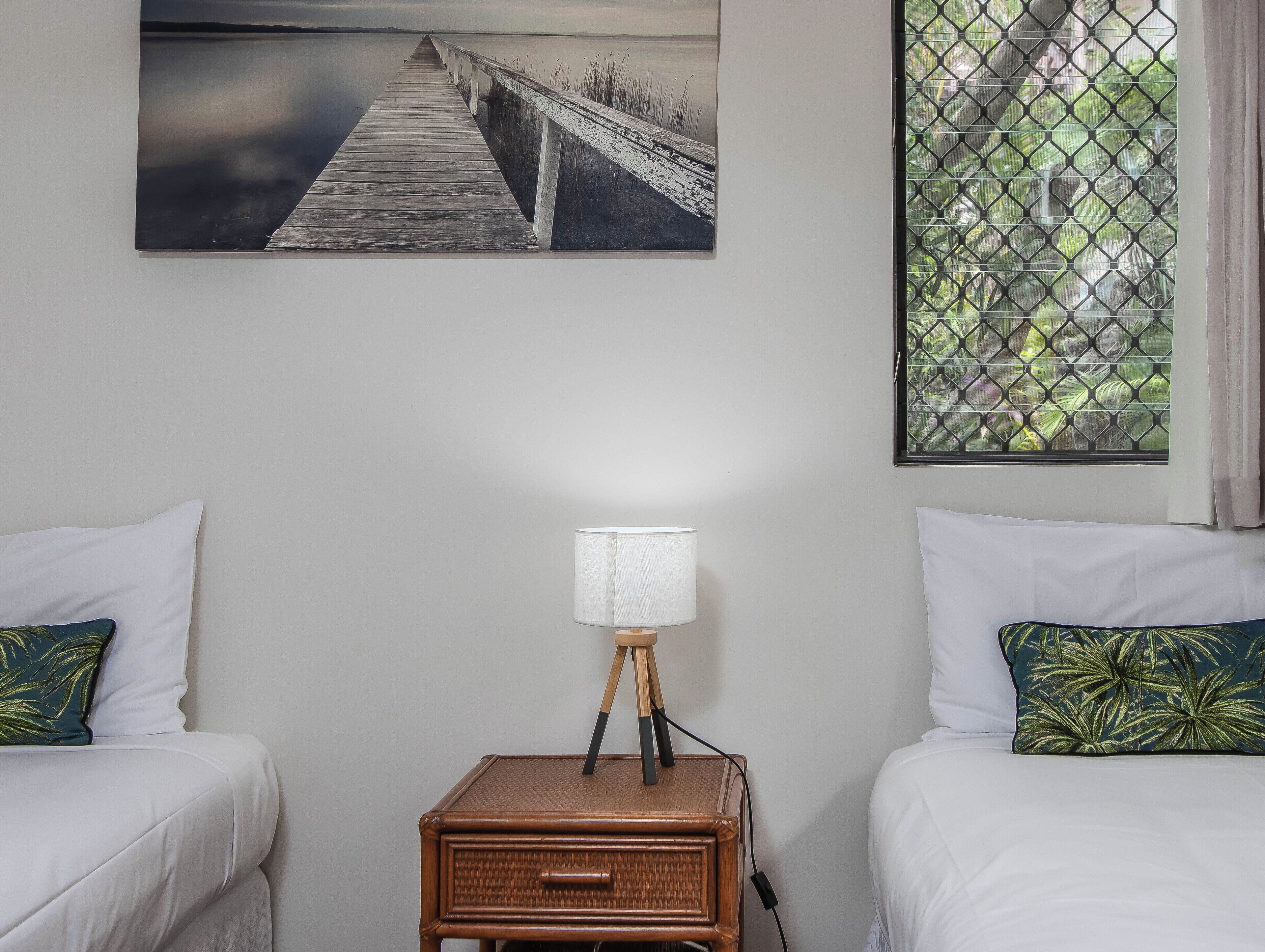 A room with two beds, a nightstand with a lamp, a picture of a pier, and a window with a view of greenery.