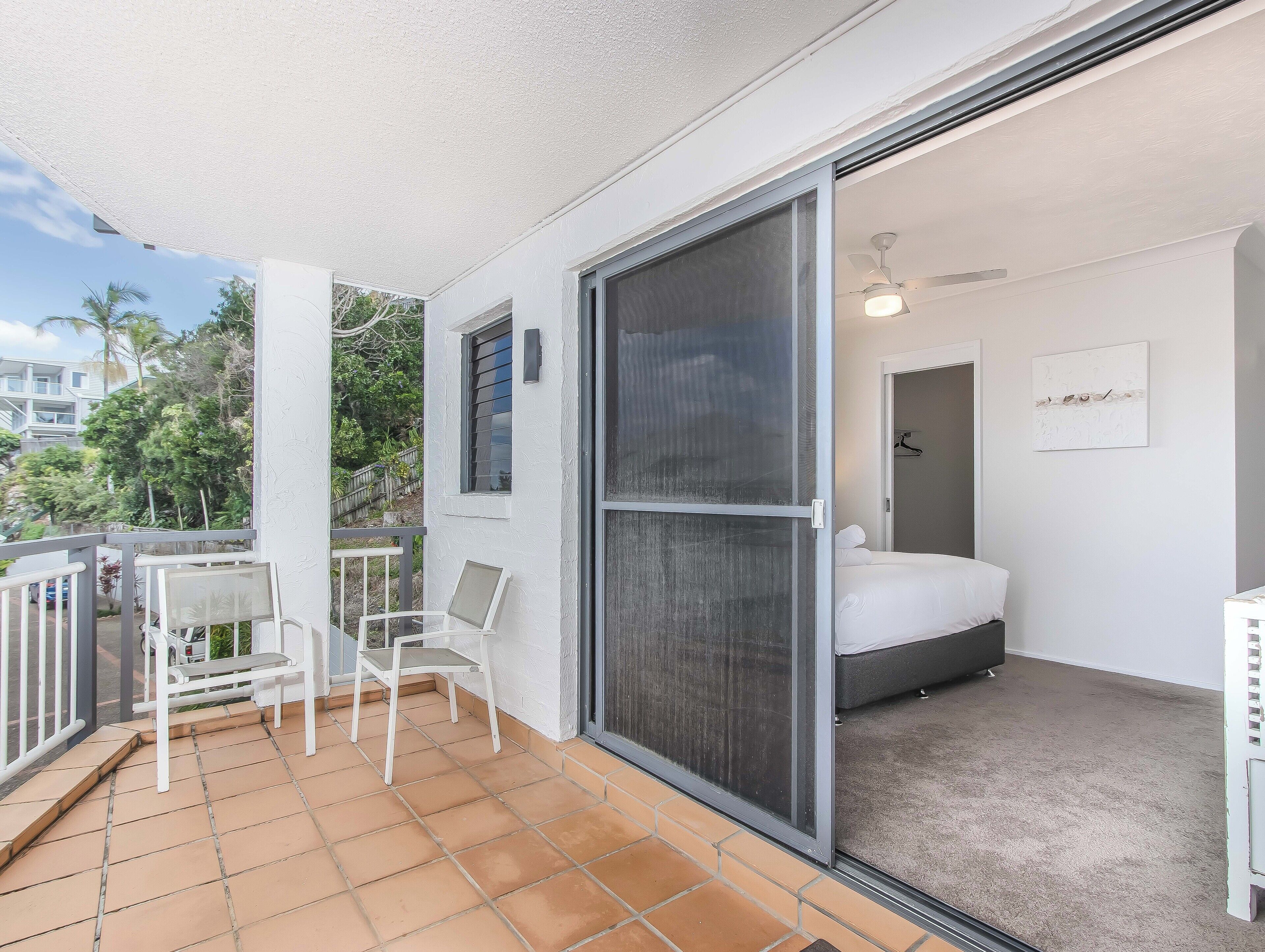 Un balcon avec une chaise blanche et une petite table, une porte-fenêtre coulissante donnant sur une chambre avec un lit et un ventilateur de plafond, et une vue sur un quartier résidentiel avec des arbres et une clôture.