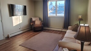 Un salon avec un fauteuil en cuir marron, un canapé beige, une table basse, une lampe et un téléviseur fixé au mur.