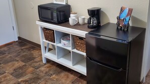 Une petite cuisine équipée d’un réfrigérateur noir, d’un four à micro-ondes, d’une machine à café et d’une petite étagère avec des paniers de rangement.