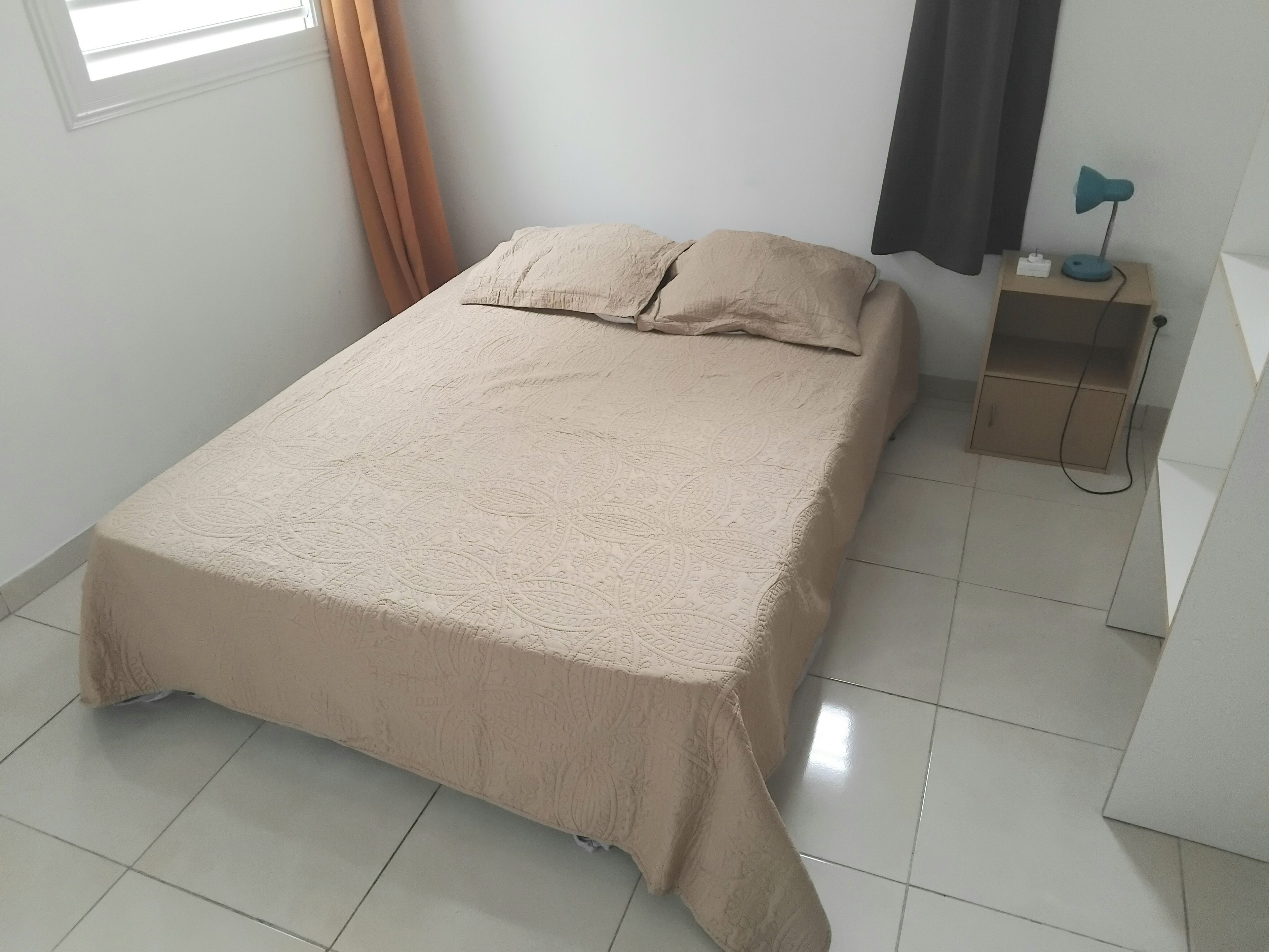 Een keurig opgemaakt bed met een beige sprei in een kamer met witte wanden, een raam met gordijnen en een nachtkastje met een lamp.
