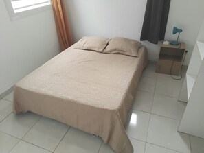 A neatly made bed with a beige bedspread in a room with white walls, a window with curtains, and a bedside table with a lamp.