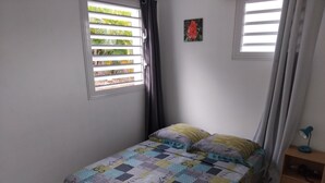 A bedroom with a bed, a window with blinds, a small table with a lamp, and a wall with a picture.