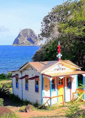 Un pequeño edificio colorido con una cruz roja en la parte superior, situado cerca de una masa de agua y una isla rocosa.